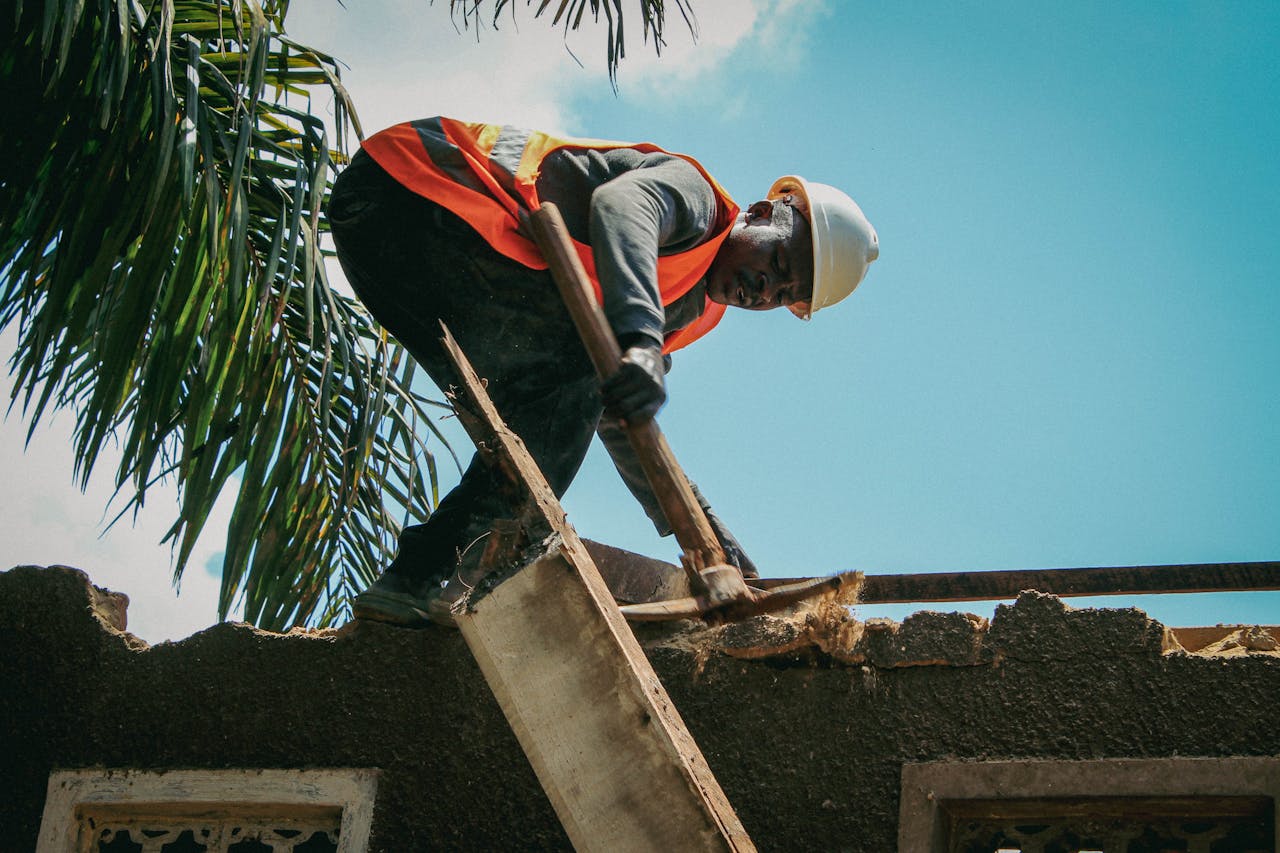 Construction worker demolishes roof under bright sky. Safety gear visible.